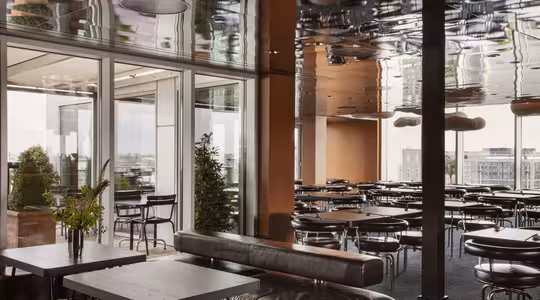 Moderne taksalong med gulv-til-tak-vinduer, reflekterende tak og svarte møbler. Rader med svarte stoler og runde bord fyller plassen, fremhevet av potteplanter og polstrede sitteplasser. Byens skyline synlig gjennom vinduene.
