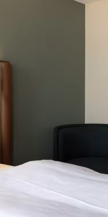 Et moderne hotellromshjørne med en seng dekket av hvitt sengetøy, en polstret brun hodegavl, et rundt beige sidebord, en mørkt trukket stol og en hvit sylindrisk vegglampe.