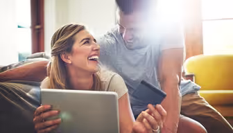 En smilende kvinne holder et nettbrett og et kredittkort, og sitter ved siden av en smilende mann. De er innendørs, med sterkt sollys som strømmer inn gjennom et vindu. Atmosfæren er munter og avslappet, og foreslår netthandel eller surfing sammen.
