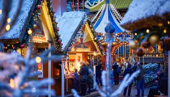 En festlig markedsscene med snødekte, dekorerte boder, varme lys og en mengde mennesker. Bodene er pyntet med julepynt, og en kaffebod er livlig med besøkende. I bakgrunnen legger fargerike strukturer til feriestemningen.