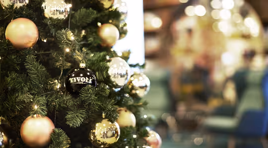 Et juletre dekorert med gull og rosa ornamenter og varme lys. En svart ornament merket "Tivoli" er fremtredende. Bakgrunnen er ute av fokus, og viser en sittegruppe med blå stoler og en reflekterende overflate.