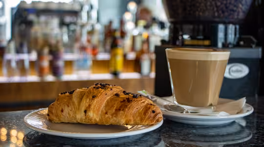 En flakete croissant med sjokoladebiter sitter på en hvit tallerken ved siden av en skummende latte i en klar glasskopp. Bakgrunnen har en uskarp visning av en kafédisk med forskjellige flasker og en kaffekvern.
