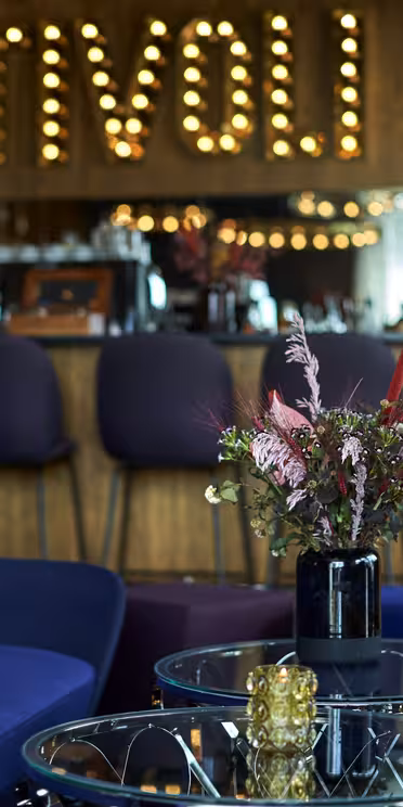 Et stilig salongområde har blå og gule sitteplasser, et bord med glassplater med blomsteroppsatser og en trebar med lilla barkrakker i bakgrunnen. Opplyste bokstaver som staver «TIVOLI» pryder veggen over baren.