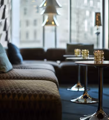 En stilig salong med vatterte sofaer, blå puter og runde trebord med dekorative lys. Rommet er dempet opplyst, med store vinduer som avslører et urbant bybilde utenfor.