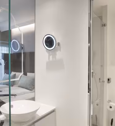 Moderne hotellrom med en glassvegg som skiller badet fra soverommet. Badet har et sirkulært opplyst speil, en hvit vask og et toalett. Soverommet har en seng med hvitt sengetøy og dempet belysning.