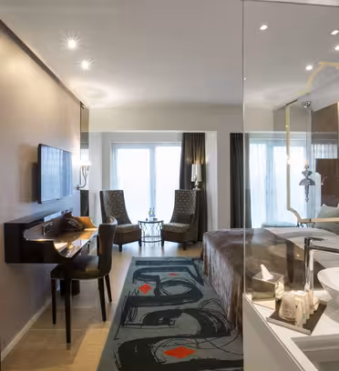 Moderne hotellrom med elegant design. Den har en seng, en veggmontert TV, et skrivebord med en stol og et sittehjørne med to mønstrede stoler ved store vinduer. Et unikt teppe og kunstneriske dekorative elementer er tilgjengelig. Vasken er synlig i det åpne rommet.
