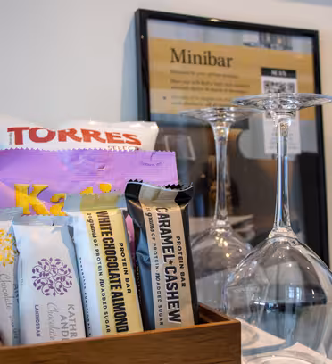 Nærbilde av et minibaroppsett med snacks som chips og sjokoladebarer, vin og glass. Et innrammet skilt viser priser på minibaren i bakgrunnen.