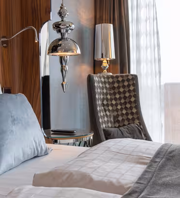 Et moderne hotellrom med en pent oppredd seng med hvitt sengetøy og lyseblå puter. Det er en stilig lampe og en elegant stol ved et vindu med utsikt over byens bygninger. Rommet har elegante tre og metalliske detaljer.