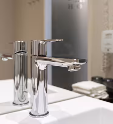 En moderne, skinnende kromkran er installert over en hvit vask på et bad. Bakgrunnen har et speil som reflekterer kranen, pent brettede håndklær og veggmonterte såpedispensere.