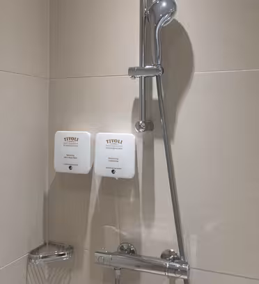 Et minimalistisk dusjoppsett med et håndholdt dusjhode i rustfritt stål på en skinne og to veggmonterte såpedispensere mot beige fliser. En liten trådkurv er festet under dispenserne for oppbevaring.
