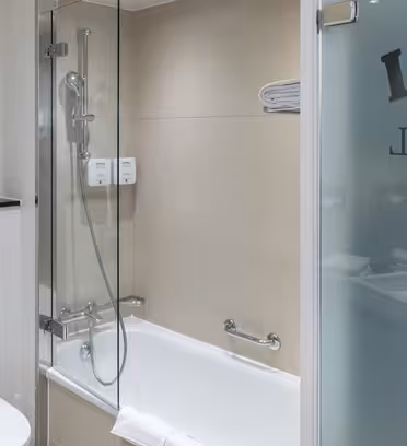 Moderne hotellbad med badekar, dusjhode og frostet glassdør. Et håndkle legges på karets kant. Beige fliser dekker veggene, og en delvis tegneseriefigur er synlig på venstre vegg.