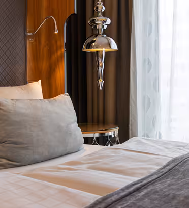 Et koselig hotellrom med en pent oppredd seng med hvitt og grått sengetøy, en stor grå pute og en moderne sølvlampe. Sengen er plassert ved siden av et vindu med rene hvite gardiner, slik at naturlig lys slipper inn i rommet.