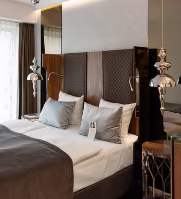 Moderne hotellrom med en stor seng med grått og hvitt sengetøy, to metalliske pendellamper på hver side og et speil bak sengegavlen. Et vindu med rene gardiner lar naturlig lys fylle rommet.