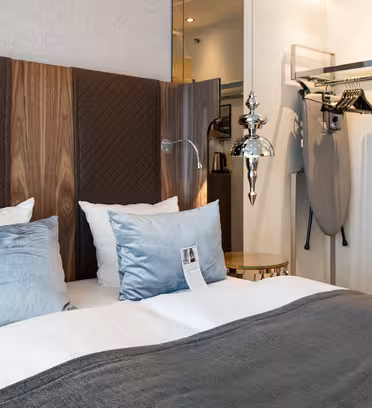 Moderne hotellrom med dobbeltseng utsmykket med hvitt og grått sengetøy, og lyseblå puter. En hodegjerde i tre har vatterte paneler. Et strykebrett, kleshengere og en vannkoker er synlige i det åpne skapet. En skinnende pendel henger over.