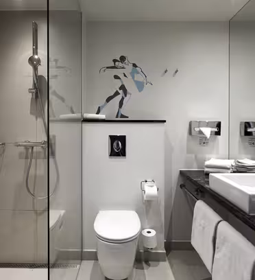 Moderne bad med dusjkabinett i glass, toalett og svart benkeplate med hvit vask. Over toalettet viser et veggdekor en stilisert figur i bevegelse. Håndklær henger under disken, og speil reflekterer det elegante designet.