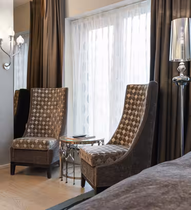 Moderne hotellrom med to mønstrede høyryggede stoler ved siden av et lite glassbord. En flatskjerm-TV er montert på veggen, og en elegant gulvlampe står i nærheten. Mørke gardiner rammer inn store vinduer med rene hvite gardiner, og skaper en koselig atmosfære.