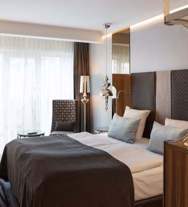 Moderne hotellrom med dobbeltseng, grått sengetøy og puter. En stilisert stol og et lite skrivebord er i nærheten av vinduet. En vegghengt TV er til venstre, og dekorative lys henger ved sengen. Et brett med fasiliteter er på treskapet. Elegant innredning.