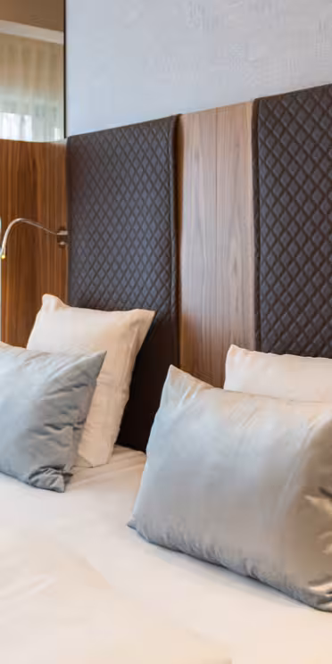 Luksuriøst hotellrom med en pent laget king-size-seng med hvitt sengetøy og grå puter. Elegante metalliske lamper er på hver side, og en plysjstol er i hjørnet. Rommet er lyst med naturlig lys fra store vinduer.