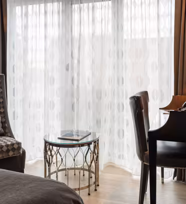 Hotellrominteriør med en høy, mønstret lenestol og et lite glassbord med magasiner nær rene gardiner. Et svart skrivebord med telefon er til høyre, supplert med en vegglampe.