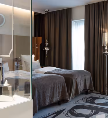 Moderne hotellrom med to enkeltsenger dekket av grå tepper. Et bad med glassvegger har en elegant hvit vask og krom-armaturer. Mørke gardiner dekker vinduene, og et mønstret teppe gir rommet et snev av eleganse.