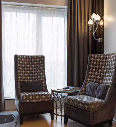 Et koselig hotellrom har to mønstrede lenestoler med mørk og lys diamantdesign, et rundt glassbord med telefon og elegante lysarmaturer. En gulvlampe og en montert TV er også synlig mot mørke gardiner.