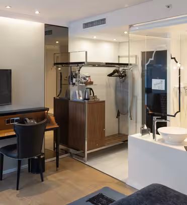 Moderne hotellrom med en stor veggmontert TV, et skrivebord med en svart stol og et åpent skap med bagasjehylle, kleshengere og kaffetrakter. En glassvegg skiller badet med vask og speil synlig.