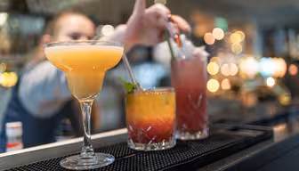 En bartender tilbereder cocktailer på en bardisk. I fokus er tre drinker: en gul margarita i et glass til venstre, en lagdelt oransje og gul drink i midten, og en rød drikke med is til høyre. Bakgrunnen er mykt uskarp.