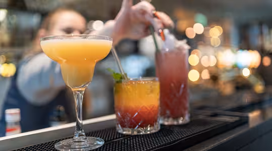 En bartender tilbereder cocktailer på en bardisk. I fokus er tre drinker: en gul margarita i et glass til venstre, en lagdelt oransje og gul drink i midten, og en rød drikke med is til høyre. Bakgrunnen er mykt uskarp.