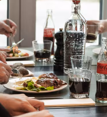 Folk nyter et måltid ved et bord med tallerkener med mat, glass og flere flasker Coca-Cola. Noen kutter mat mens andre skjenker på drikke. Innstillingen fremstår uformell og avslappet, med naturlig lys som kommer gjennom et nærliggende vindu.