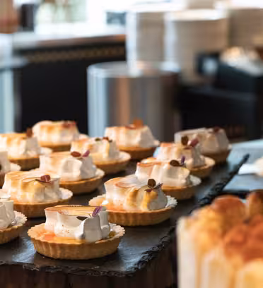 En visning av diverse desserter på et bord, med sitronmarengsterter toppet med små spiselige dekorasjoner. I bakgrunnen er det flere desserter, tallerkener og myk dekor.