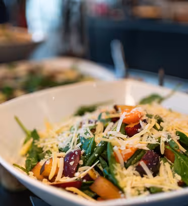 En hvit bolle fylt med en fargerik salat med fersk spinat, oppskåret rødløk, paprika og en sjenerøs topping av revet ost. Salaten er satt på en reflekterende mørk overflate med uskarpe retter i bakgrunnen.