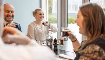Fire personer sitter ved et bord i et lyst rom, smiler og holder drinker. En rekke drikkevarer, inkludert en flaske vann og en Coca-Cola, står på bordet. Atmosfæren fremstår som avslappet og vennlig.