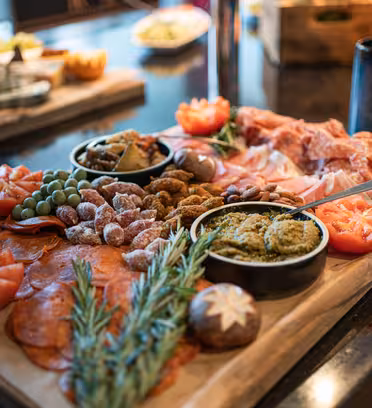 Et charcuteribrett med oppskåret kjøtt, nøtter og grønnsaker. Den inkluderer tre tomatblomster, rosmarin og separate skåler med dipper. Et glass og diverse matvarer er uskarpe i bakgrunnen på et spisebord.