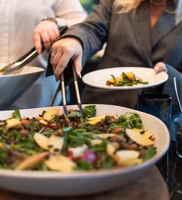 To personer som serverer seg selv fra en buffet. En stor salatskål inneholder grønt, epleskiver og nøtter. En person bruker en tang og holder en tallerken med salat. En liten krukke med frø og en pepperkvern er synlig i nærheten.