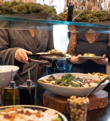 Folk serverer seg selv fra buffet med ulike salater og dressinger i hvite boller. En glasshylle over viser dekorative planter. Bakgrunnen viser flere mennesker og et godt opplyst interiør.