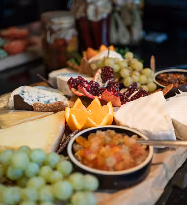 Et gourmetostfat med forskjellige oster, skivede appelsiner, halve granateple, druer og boller med chutney. Bakgrunnen viser en svak visning av mer kuttet frukt og grønt.