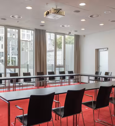 Et moderne konferanserom med U-formet bord og svarte stoler, satt mot vinduer med beige gardiner. Rommet har en rød løper og en takmontert projektor.