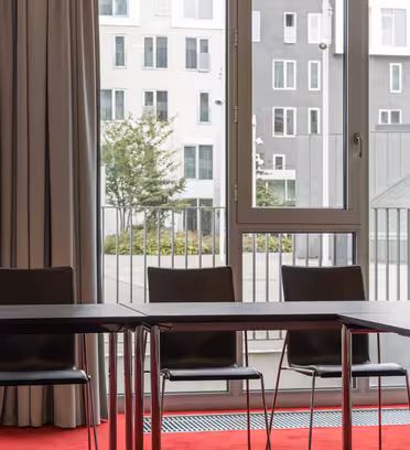Et konferanserom med tomme svarte stoler og et rektangulært bord med mørk overflate. Store vinduer viser en urban scene med moderne bygninger og grøntområder utenfor. Beige gardiner rammer inn vinduene, og et rødt teppe dekker gulvet.