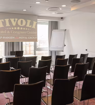 Et lite konferanserom med rader med tomme svarte stoler vendt mot en projeksjonsskjerm. Skjermen viser "TIVOLI Hotel & Congress Center." Det er en flipover-stativ og et vindu med lys som strømmer inn.