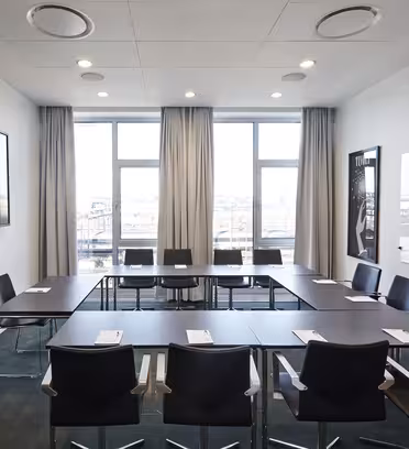 Et moderne konferanserom med et rektangulært bord og ti sorte stoler. Det er en stor TV-skjerm på veggen, en tavle og vinduer med beige gardiner som slipper inn naturlig lys. Notisblokker og penner legges på bordet.