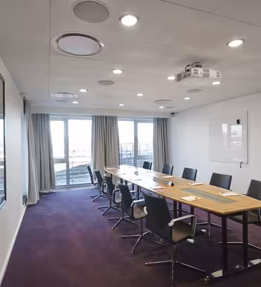 Et moderne konferanserom med et langt trebord og svarte stoler. Rommet har projektor i taket, tavle på veggen, og store vinduer med gardiner som slipper inn naturlig lys. Gulvet er teppebelagt i mørk lilla.