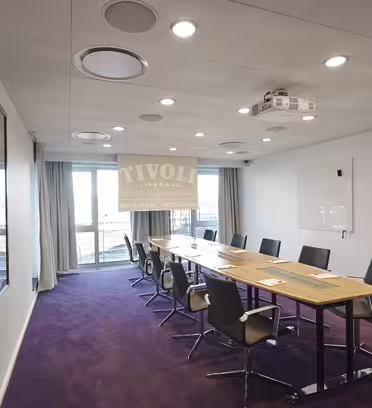 Et moderne konferanserom har et langt trebord omgitt av svarte stoler på et lilla teppe. Store vinduer ytterst lar naturlig lys fylle plassen, og en projektor henger i taket. En tavle er montert på høyre vegg.