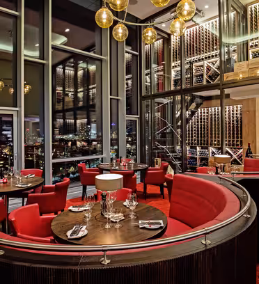 Luksuriøst restaurantinteriør med runde trebord og røde stoler. Plassen har høye vinreoler, store vinduer med utsikt over byen og elegante hengende lys som skaper en sofistikert og innbydende atmosfære.