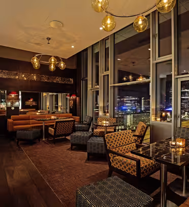 Elegant restaurantinteriør med dempet belysning. Har mønstrede stoler, plysjkabiner og marmorbord. Store vinduer gir utsikt over byen om natten. Hengende sfæriske lys gir varme til den moderne innredningen.