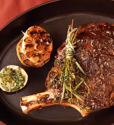 En tilberedt rib-eye biff garnert med rosmarin presenteres i en svart oval tallerken. Til biffen følger en stekt hvitløk og en halv sitron toppet med urter. Fatet hviler på en rød vattert overflate.