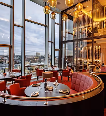Luksuriøs restaurantinteriør med gulv-til-tak-vinduer med panoramautsikt over byen. Har røde, runde boder, trebord, gylne lysarmaturer og en vinvegg. Dagtid, skyet himmel synlig utenfor.