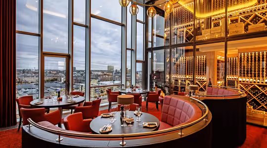 Luksuriøs restaurantinteriør med gulv-til-tak-vinduer med panoramautsikt over byen. Har røde, runde boder, trebord, gylne lysarmaturer og en vinvegg. Dagtid, skyet himmel synlig utenfor.