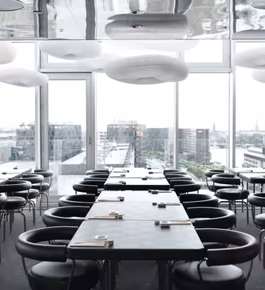 Moderne restaurantinteriør med elegante svarte bord og stoler arrangert i rader. Store vinduer gir utsikt over byen. Taket har hvite, skylignende lysarmaturer som skaper en luftig atmosfære. Naturlig lys fyller rommet.