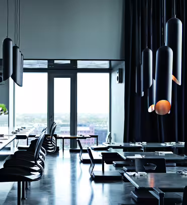 Moderne restaurantinteriør med elegante svarte bord og stoler. Unike sylindriske pendellamper henger fra taket. Store vinduer gir rikelig med naturlig lys og utsikt over bybildet. Minimalistisk og stilig design.