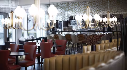 Et moderne restaurantinteriør med røde og beige sitteplasser, mørke runde bord og en elegant bar i bakgrunnen. Utsmykkede gulllysekroner henger fra taket og gir en varm glød. Veggene har en geometrisk mønstret dekor.